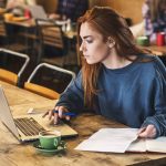 Young woman with long red hair sitting at table, working on laptop computer.